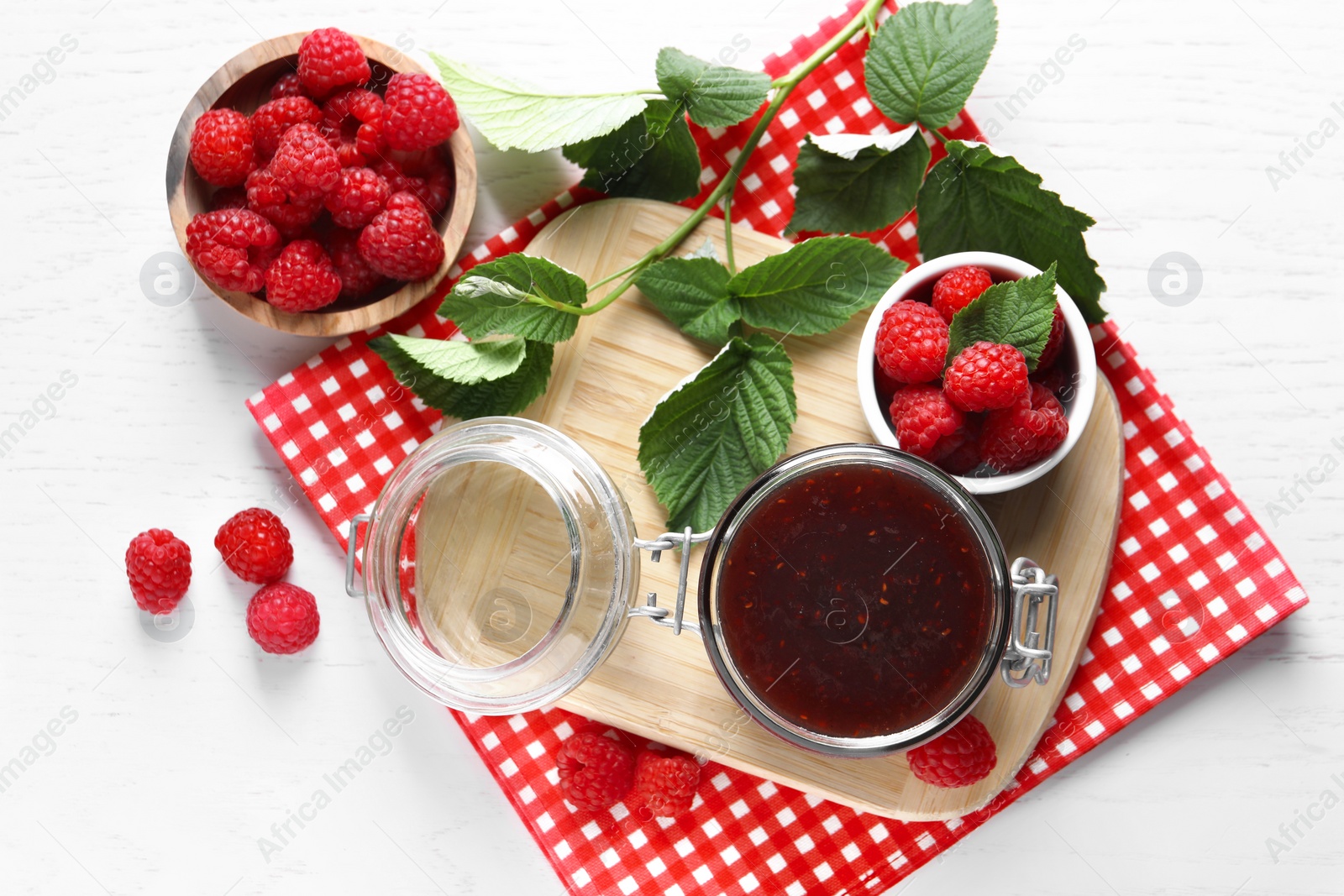 Photo of Jar of delicious raspberry jam, fresh berries and green leaves on white wooden table, flat lay