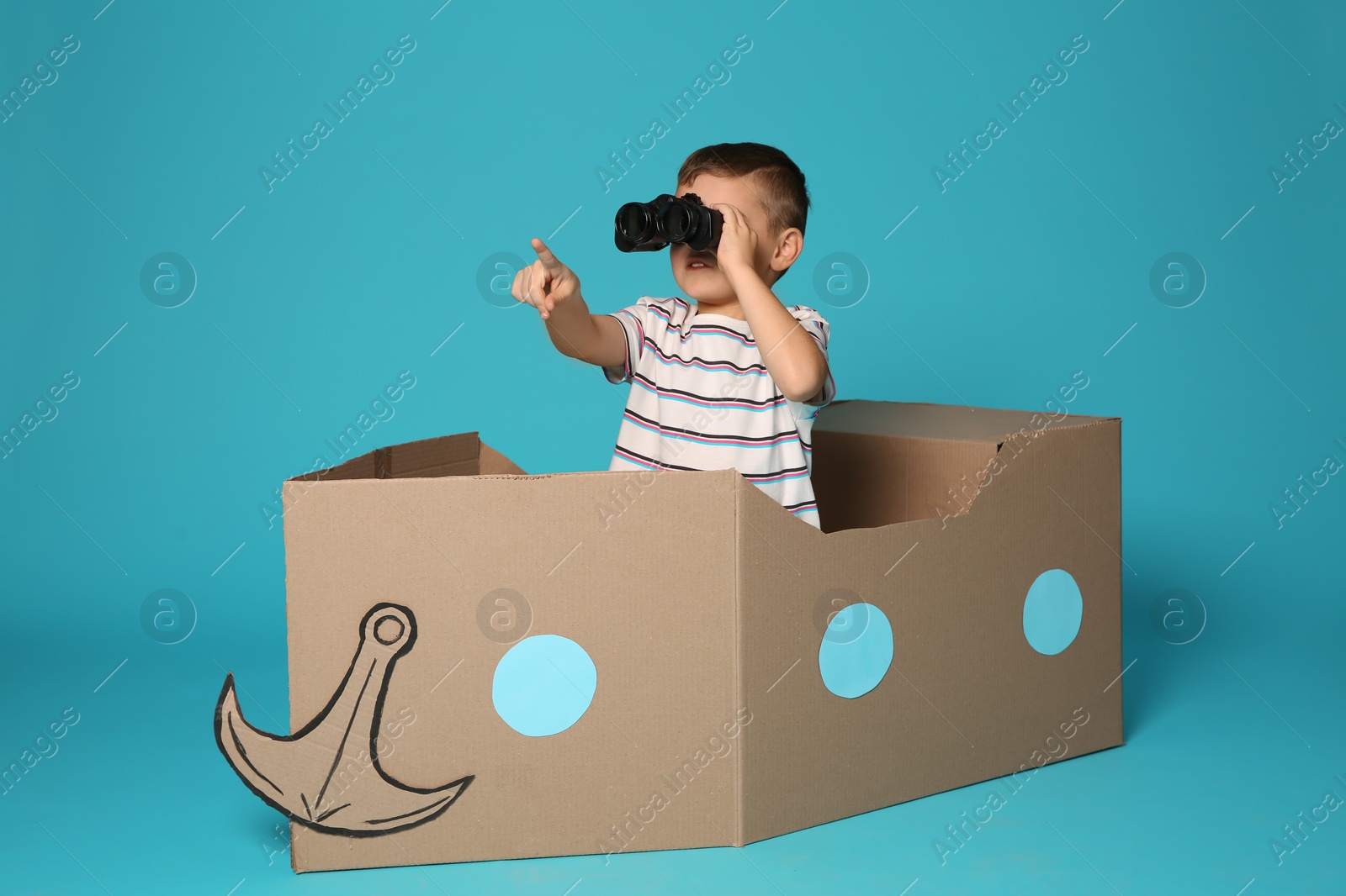 Photo of Cute little boy playing with binoculars and cardboard boat on color background
