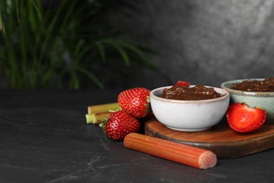 Tasty rhubarb jam in bowls, cut stems and strawberries on dark textured table. Space for text