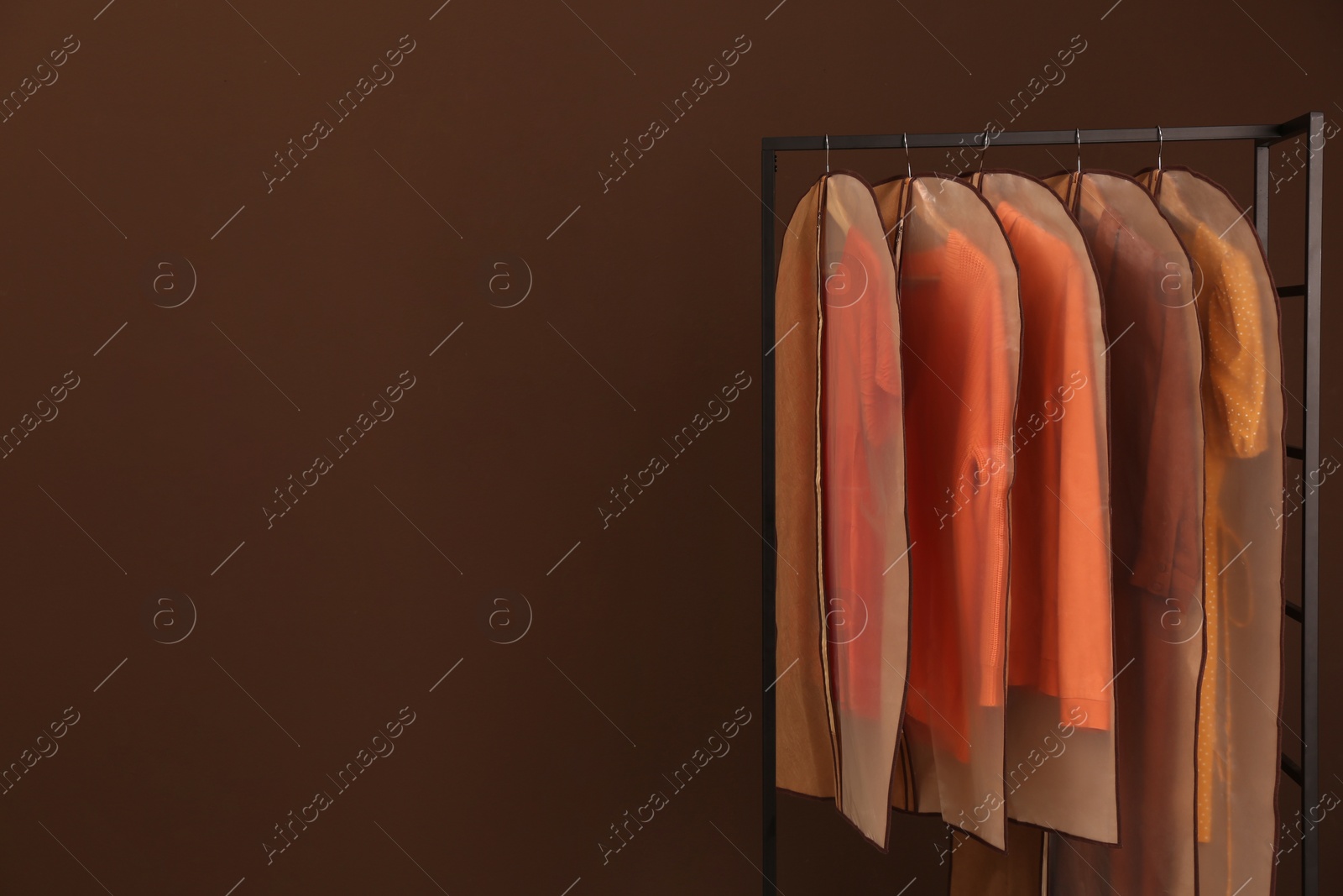 Photo of Garment bags with clothes on rack against brown background. Space for text