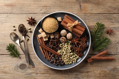 Plate with different aromatic spices, spoons and fir branches on wooden table, flat lay