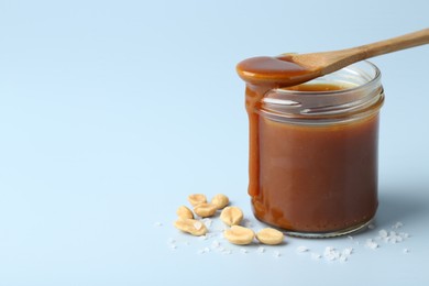 Photo of Jar of tasty salted caramel, spoon and peanuts on light blue background, closeup. Space for text