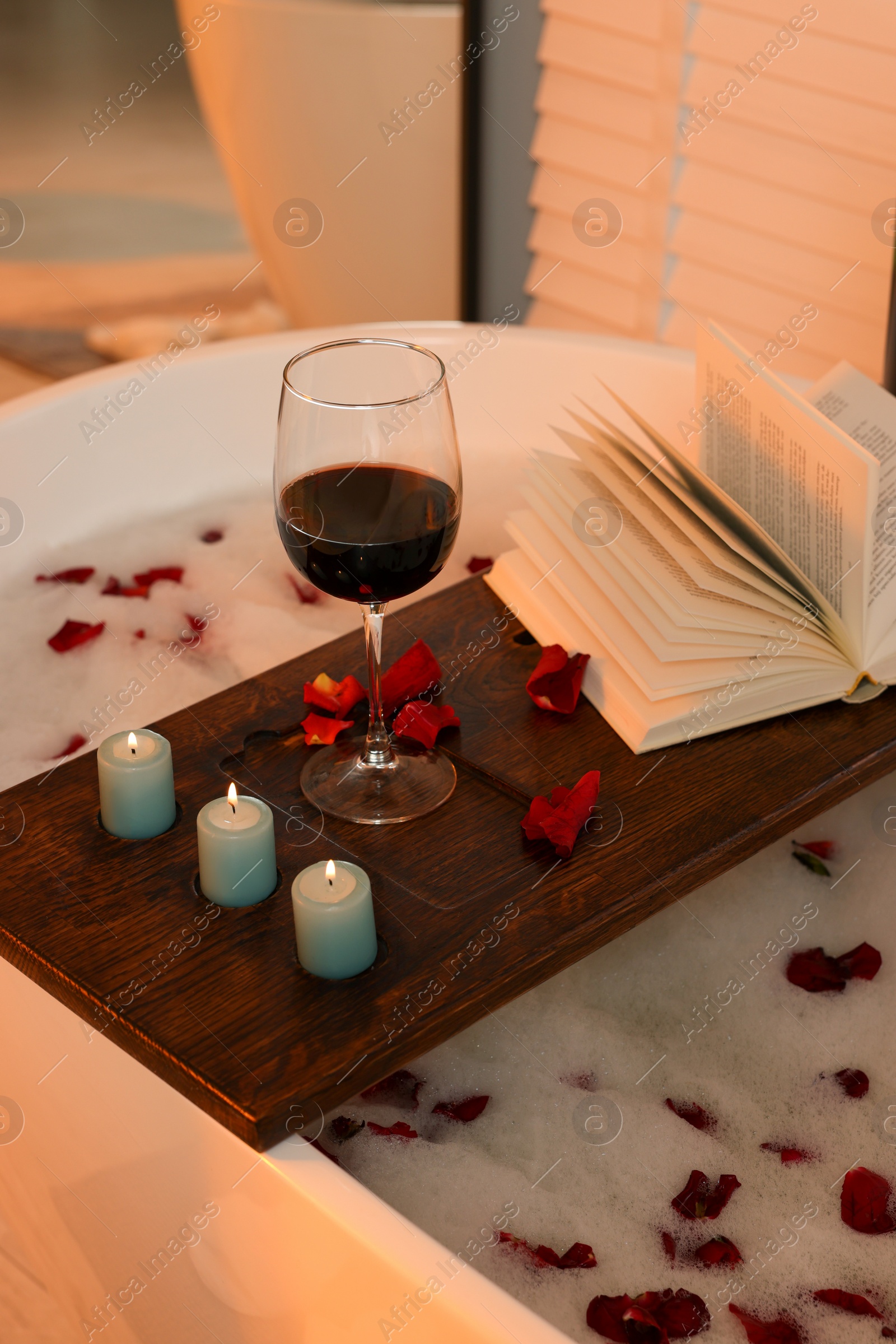 Photo of Wooden board with glass of wine, book, burning candles and rose petals on bath tub