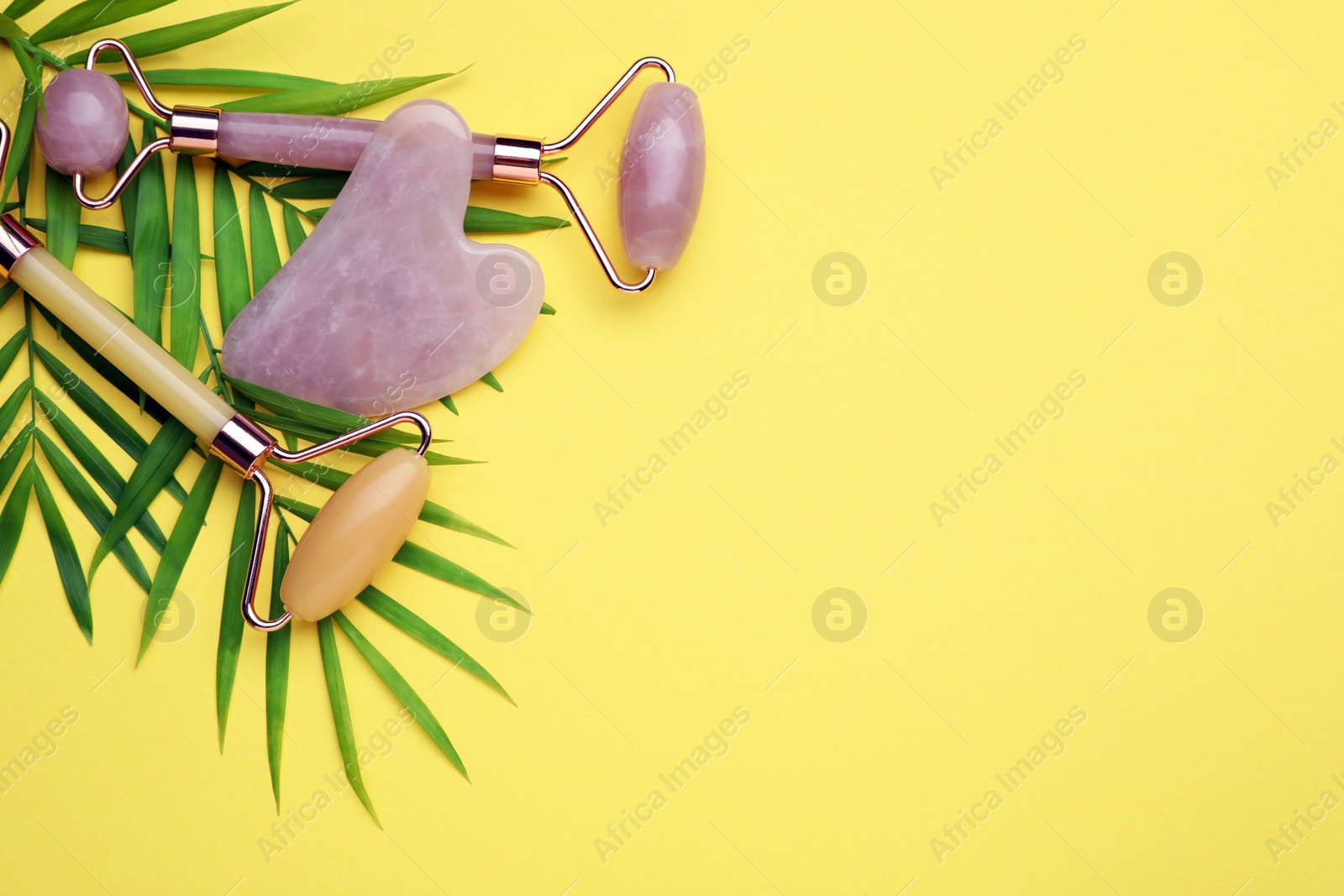 Photo of Gua sha stone, different face rollers and green leaves on yellow background, flat lay. Space for text