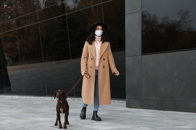 Photo of Woman in protective mask with German Shorthaired Pointer on city street. Walking dog during COVID-19 pandemic
