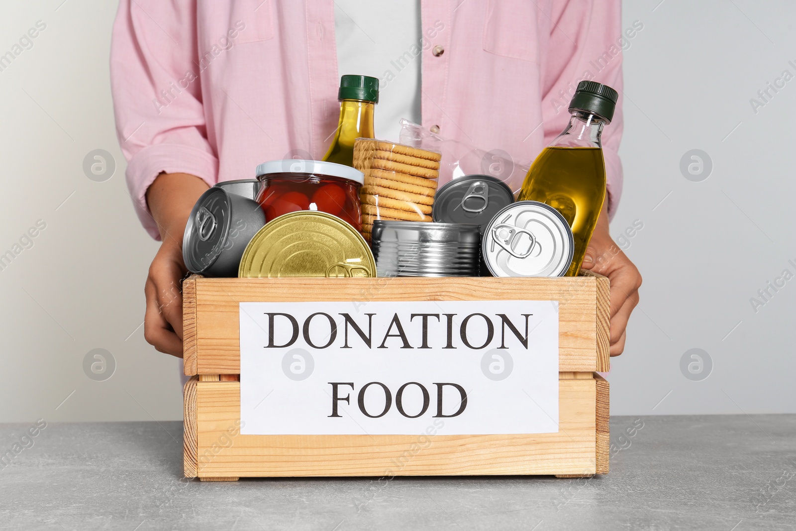 Photo of Woman holding donation crate with food at light grey table, closeup