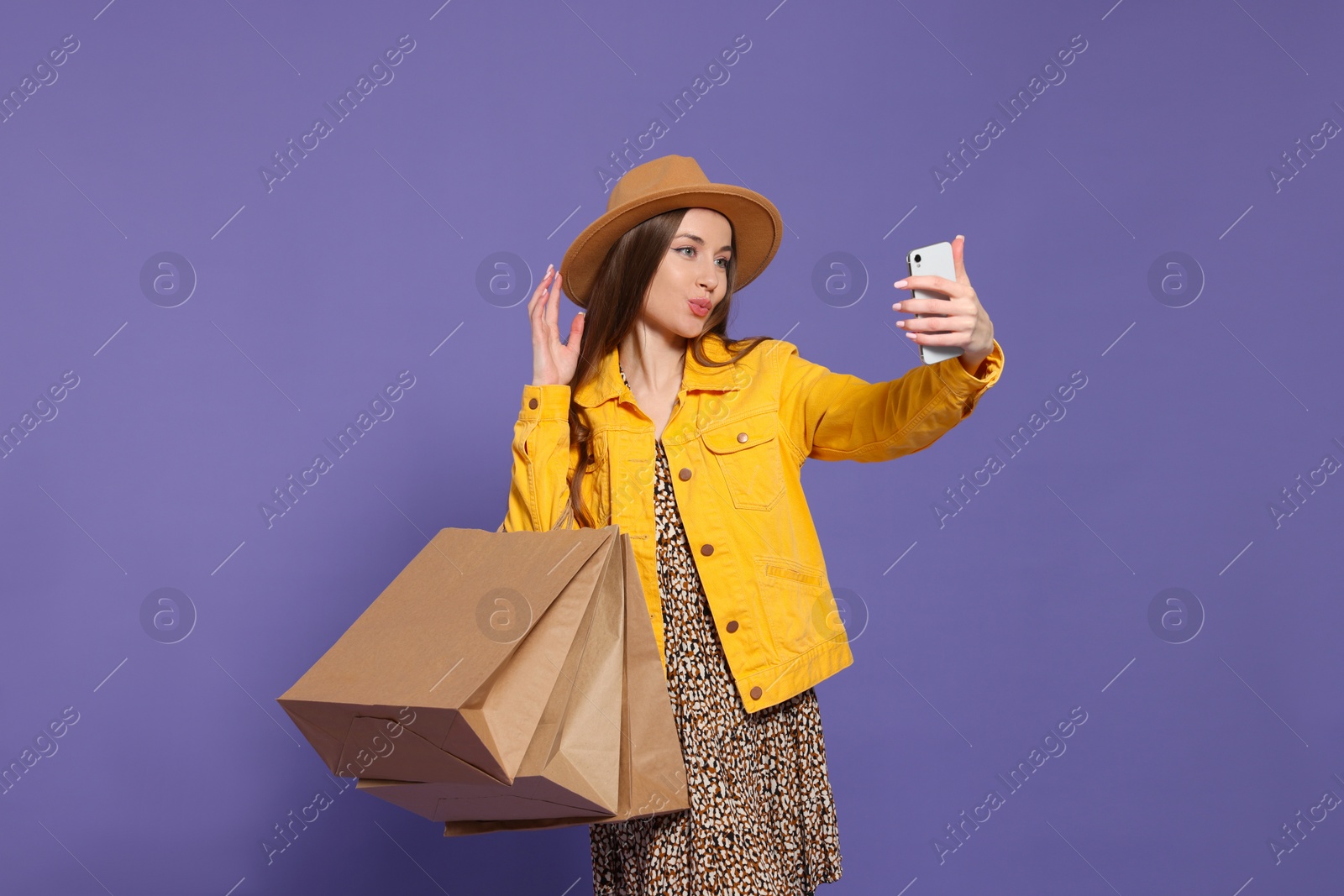 Photo of Stylish young woman with shopping bags taking selfie on purple background