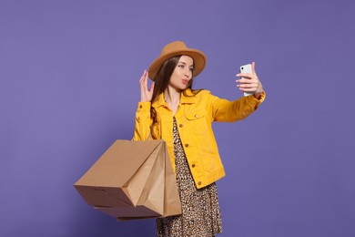 Stylish young woman with shopping bags taking selfie on purple background