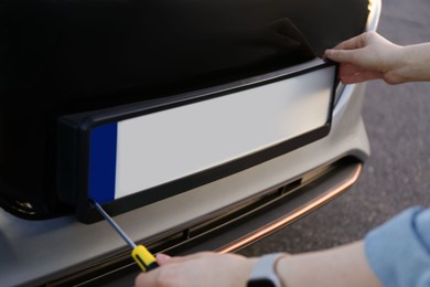 Woman with screwdriver installing vehicle registration plate to car outdoors, closeup