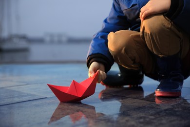Photo of Little boy playing with paper boat near puddle on pier, closeup