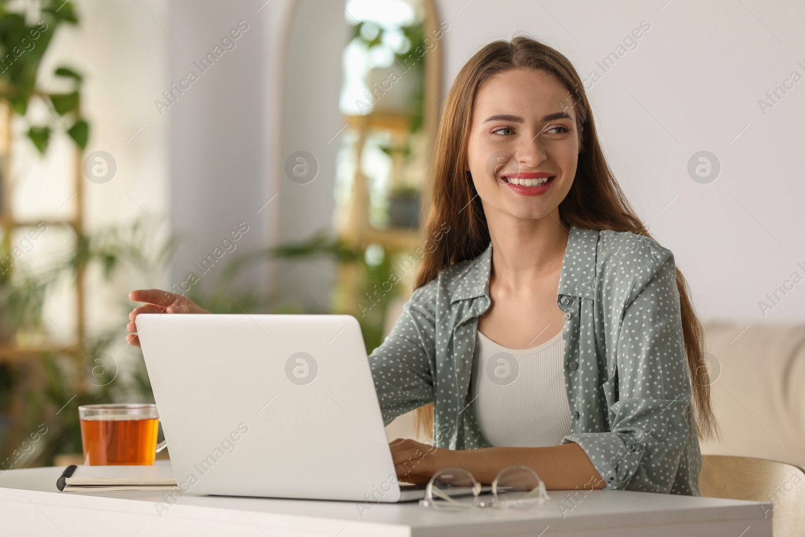 Photo of Happy young woman with laptop and tea at table indoors