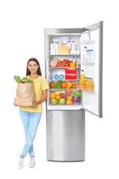 Young woman with bag of groceries near open refrigerator on white background