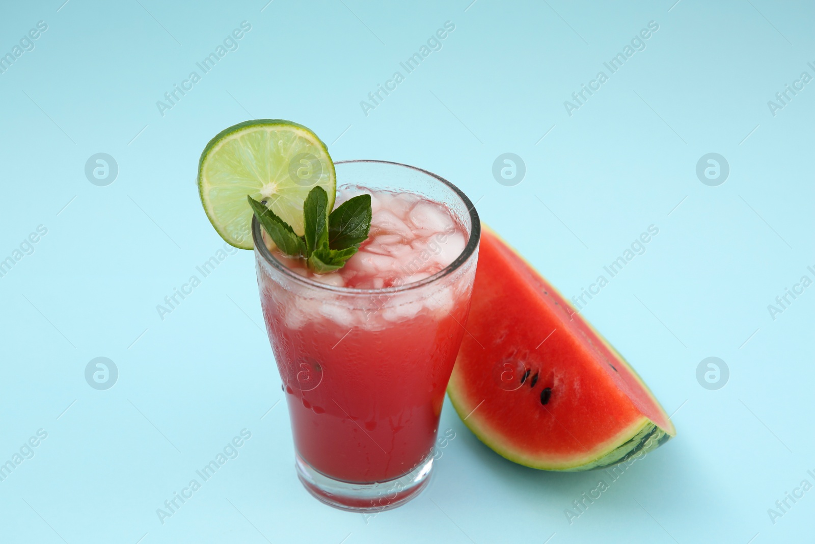 Photo of Glass of delicious drink with lime, ice cubes and cut fresh watermelon on light blue background