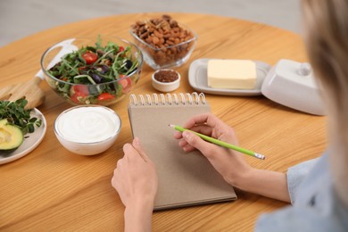 Woman with notebook and different products at wooden table, closeup. Keto diet