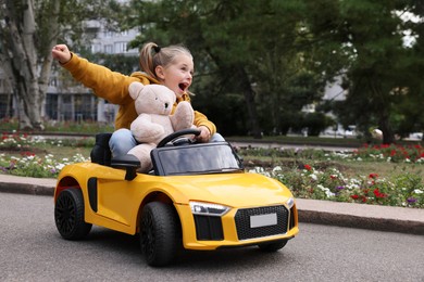 Photo of Cute little girl with toy bear driving children's car in park. Space for text