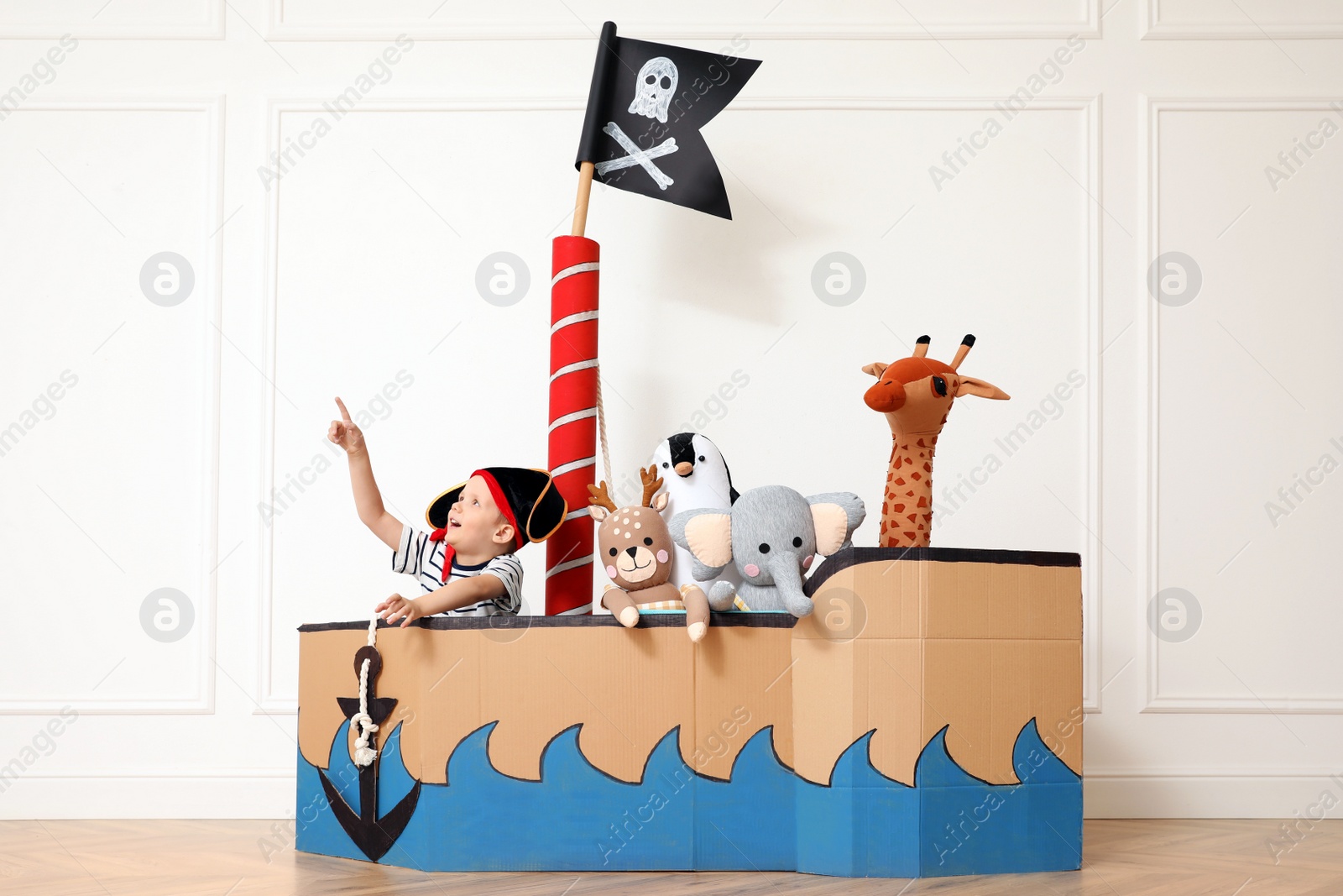 Photo of Cute little boy playing with toys in pirate cardboard ship near white wall indoors