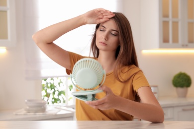 Photo of Woman with portable fan at table in kitchen. Summer heat