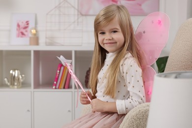 Photo of Cute little girl in fairy costume with pink wings and magic wand in armchair at home, space for text