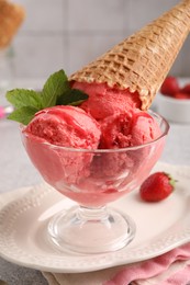 Photo of Delicious scoops of strawberry ice cream with mint and wafer cone in glass dessert bowl served on grey table, closeup