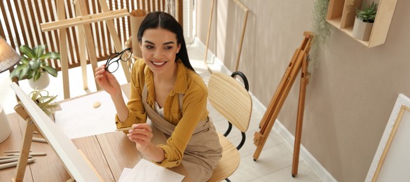 Image of Young woman drawing on easel with pencil at table indoors, above view. Banner design
