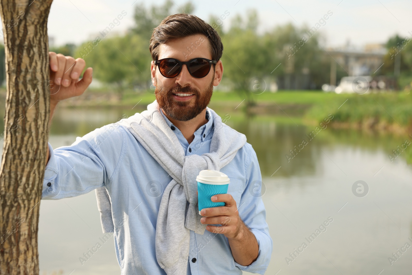 Photo of Portrait of handsome bearded man in sunglasses with coffee near lake, space for text