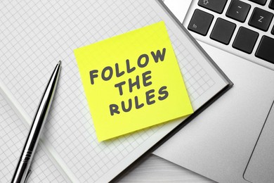 Image of Paper note with phrase Follow The Rules, pen and laptop on wooden table, flat lay