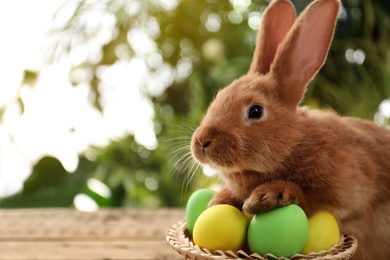 Photo of Cute bunny and basket with Easter eggs on table against blurred background. Space for text