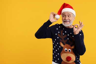 Photo of Senior man in Christmas sweater and Santa hat showing OK gesture on orange background. Space for text