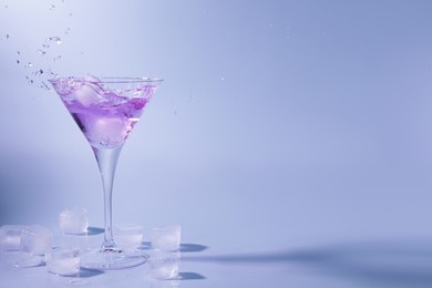 Photo of Cocktail splashing out of martini glass near ice cubes on light blue background, space for text