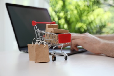 Photo of Internet shopping. Small cart with boxes and bags near man using laptop indoors