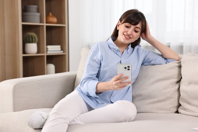 Photo of Beautiful young housewife using smartphone on sofa at home
