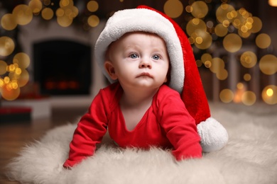 Photo of Cute little baby in red pajamas and Santa hat on floor against blurred festive lights. Christmas suit