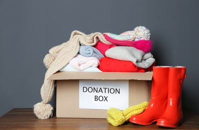 Photo of Donation box with knitted clothes and rubber boots on table near grey wall