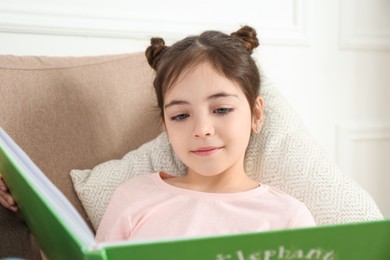 Photo of Little girl reading book in armchair at home