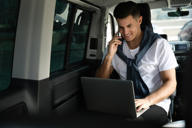 Photo of Handsome man with laptop talking on phone in modern car