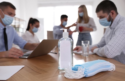 Photo of Protective masks, hand sanitizer and blurred view of coworkers on background. Business meeting during COVID-19 pandemic