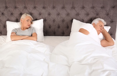 Photo of Upset mature woman lying in bed near her sleeping husband at home. Relationship problems