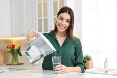 Photo of Woman pouring water from filter jug into glass in kitchen