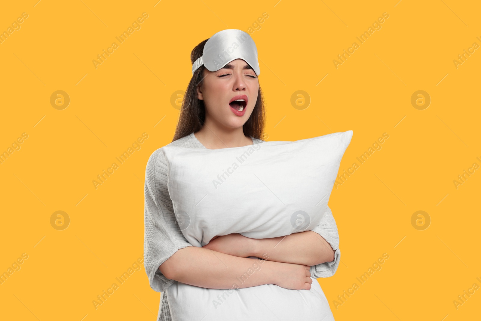 Photo of Tired young woman with sleep mask and pillow yawning on yellow background. Insomnia problem