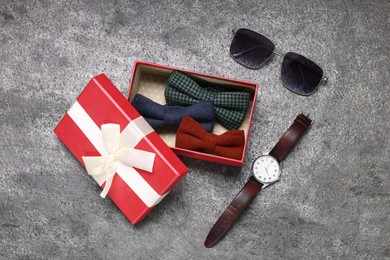 Photo of Stylish color bow ties in gift box, wristwatch and sunglasses on grey textured background, flat lay