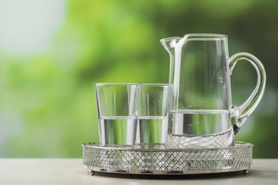 Photo of Jug and glasses with clear water on white table against blurred green background, closeup. Space for text