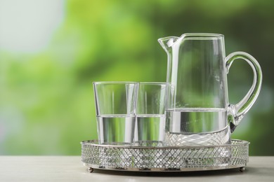 Jug and glasses with clear water on white table against blurred green background, closeup. Space for text