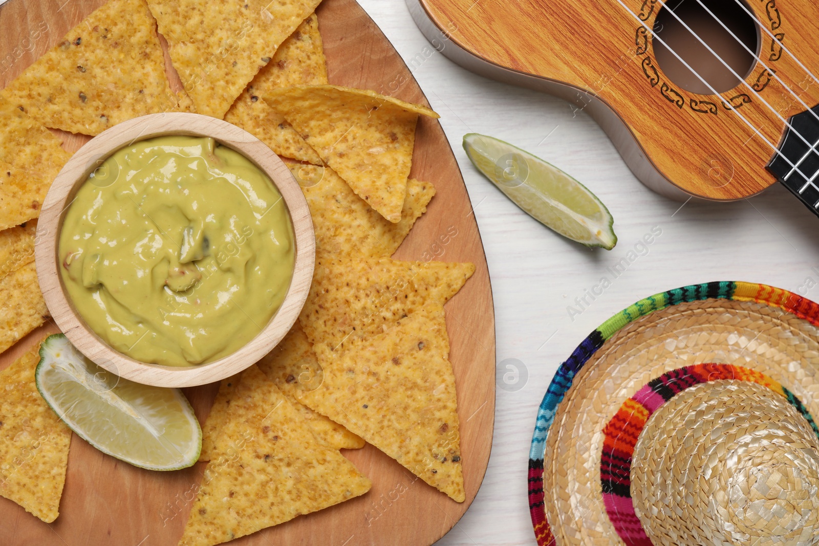 Photo of Delicious guacamole with nachos chips, Mexican sombrero hat and ukulele on white wooden table, flat lay