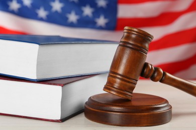 Photo of Judge's gavel and books on white wooden table against American flag, closeup