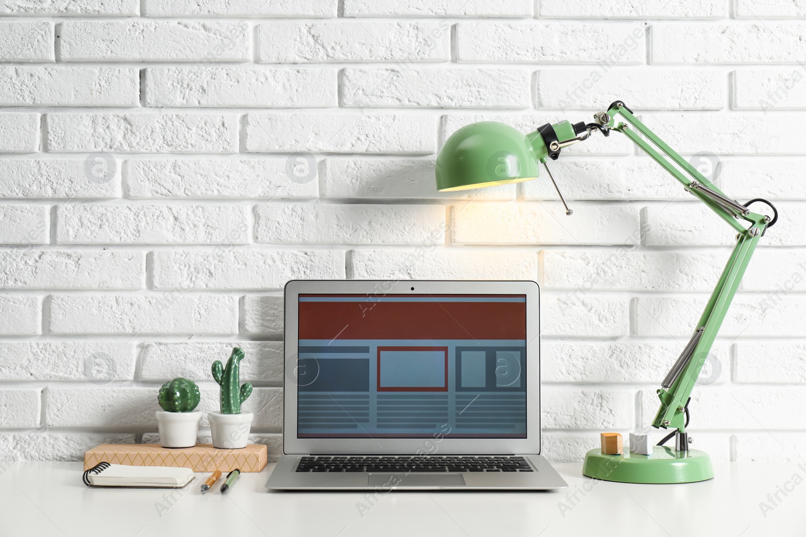 Photo of Modern workplace with laptop and lamp on table near brick wall. Space for design