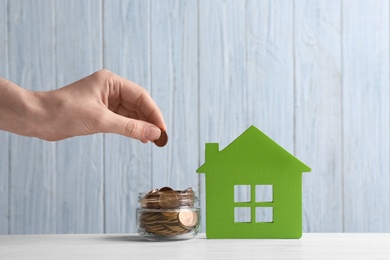 Woman putting coin into jar near house model on table against wooden background. Space for text