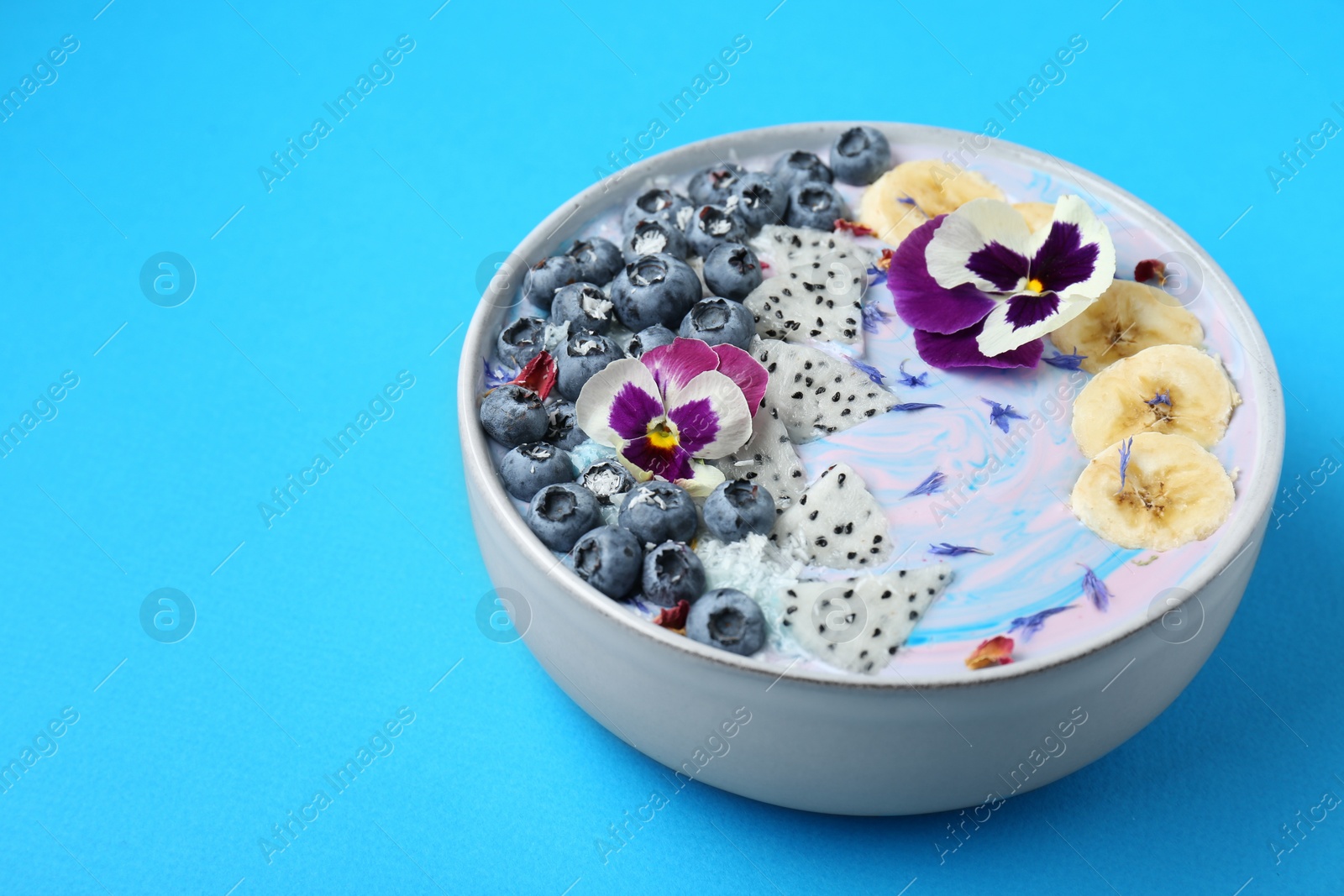 Photo of Delicious smoothie bowl with fresh fruits, blueberries and flowers on light blue background, closeup. Space for text