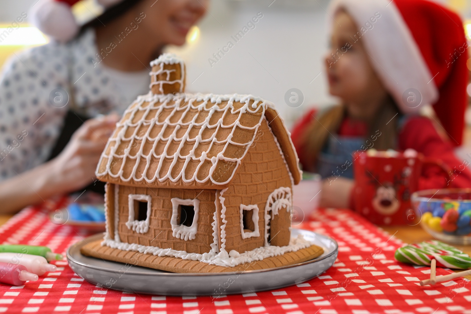 Photo of Mother and daughter in Santa hats cooking together, focus on gingerbread house