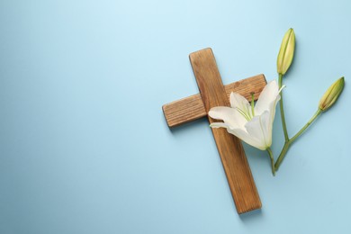 Photo of Wooden cross and lily flowers on light blue background, top view with space for text. Easter attributes
