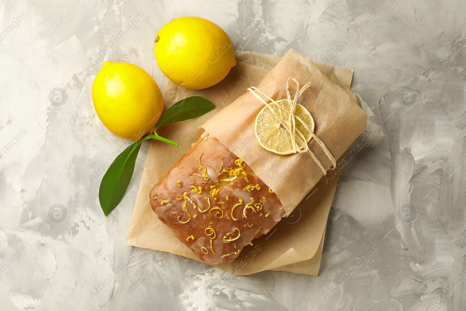 Photo of Wrapped tasty lemon cake with glaze and citrus fruits on light grey textured table, top view